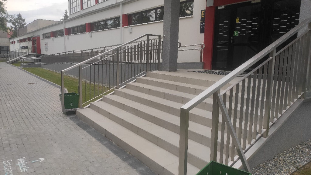 Widok z zewnątrz. Po prawej stronie widać schody z poręczami i wejście do budynku. Po lewej chodnik z białym napisem Wejście Entrance i strzałką.