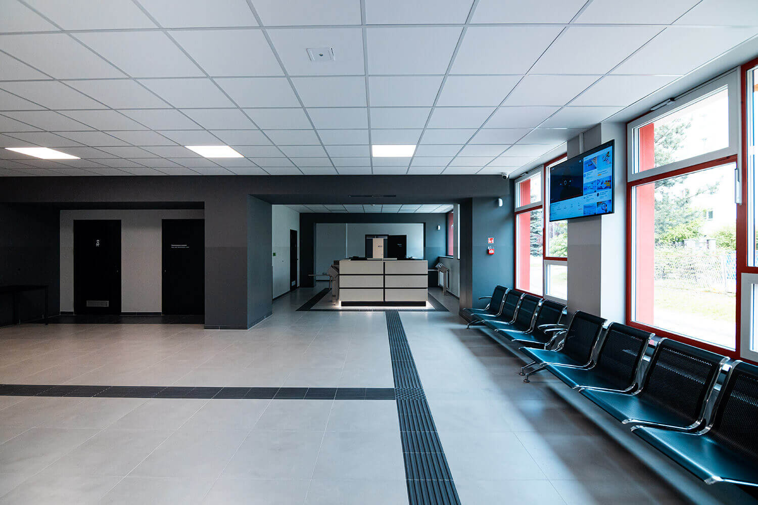 Wnętrze budynku, widok na kasę i bramkę wpuszczającą na basen. Po prawej stronie widać okna i telewizor wiszący na ścianie, po lewej korytarz i dwoje drzwi prowadzących do pomieszczeń. Na podłodze ścieżki naprowadzające.